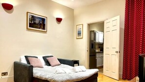 A bedroom with a bed, a small table, and a door leading to a kitchen. - Birmingham Cottage Contractors Family SelfCheck in (West Midlands)