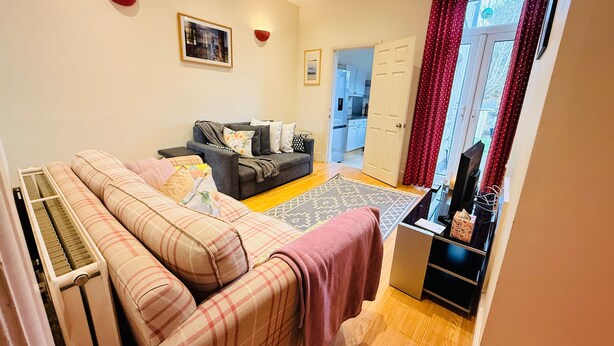 A living room with a sofa, a coffee table, and a TV stand. There is a door leading to a kitchen and a window with curtains. - Birmingham Cottage Contractors Family SelfCheck in (West Midlands)