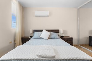 A bedroom with a bed, two bedside tables, a nightstand, a window with curtains, and an air conditioning unit on the wall. - Tropical Sunset Penthouse • Rooftop & Hot Tub (Cabo Rojo)