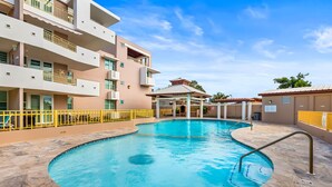 A swimming pool with a tiled deck, a covered seating area, and a multi-story building with balconies. - Tropical Sunset Penthouse • Rooftop & Hot Tub (Cabo Rojo)