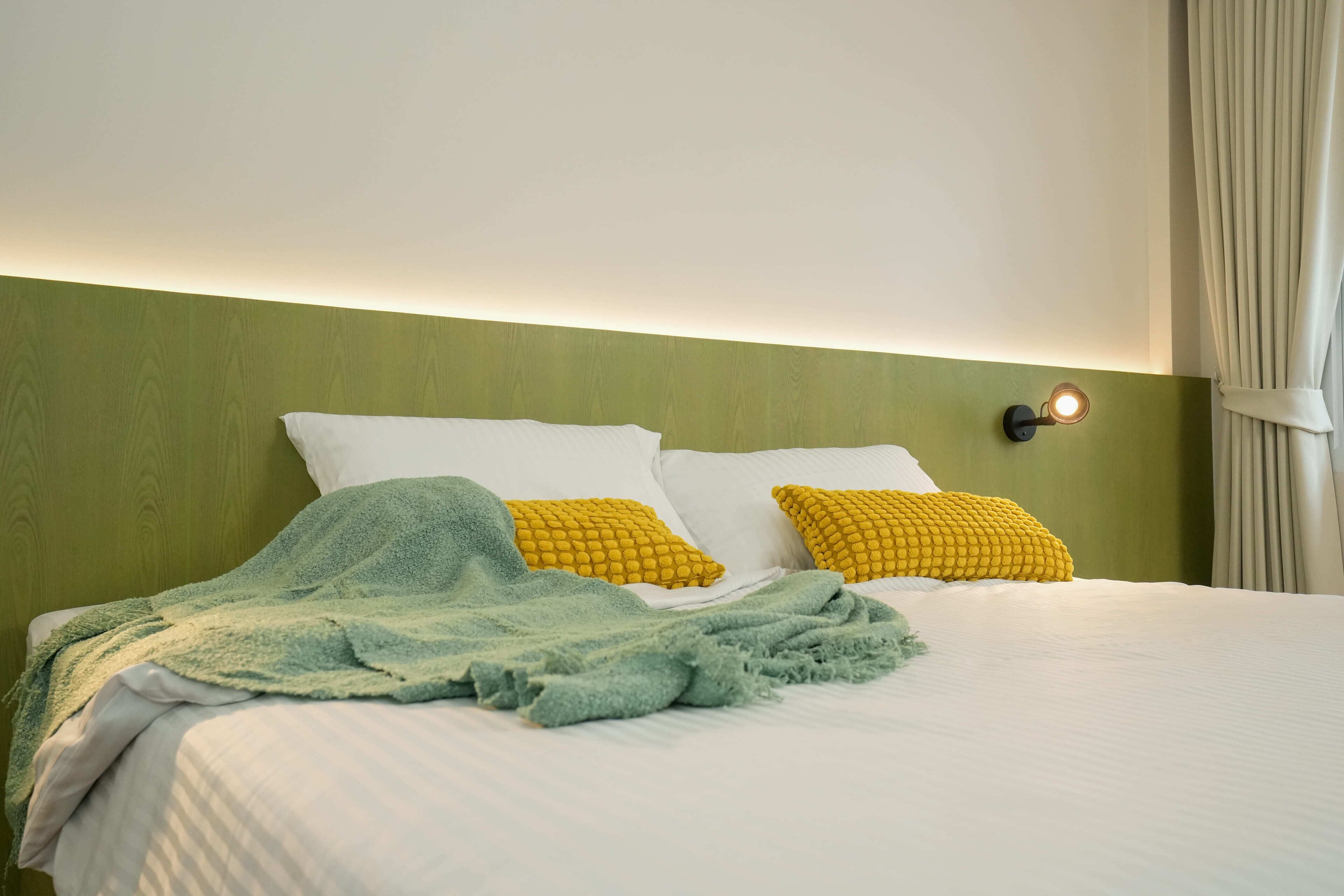 A neatly made bed with white and yellow pillows, a green blanket, and a wall-mounted light fixture.