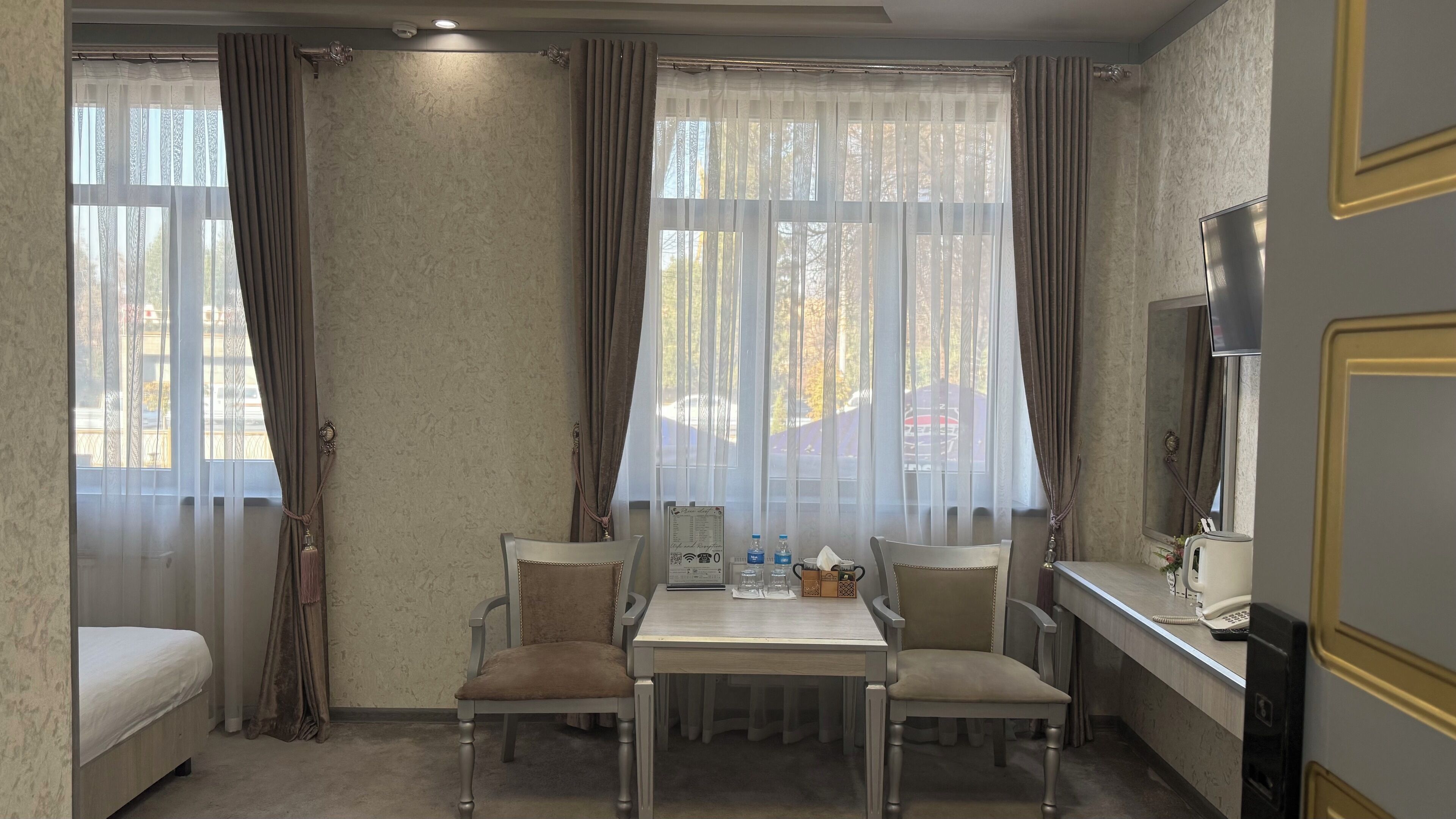 A hotel room with a bed, a desk with a telephone, two chairs, a table, and a large window with curtains.