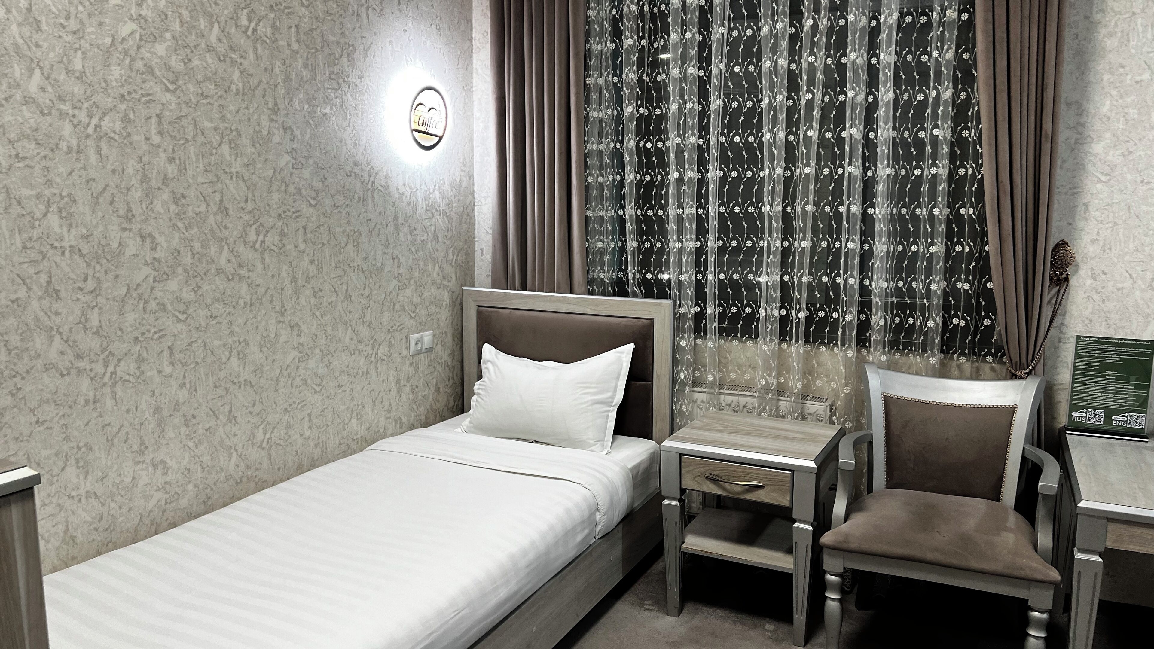 A single bed with a headboard, a bedside table, a chair, and a small desk with a chair.