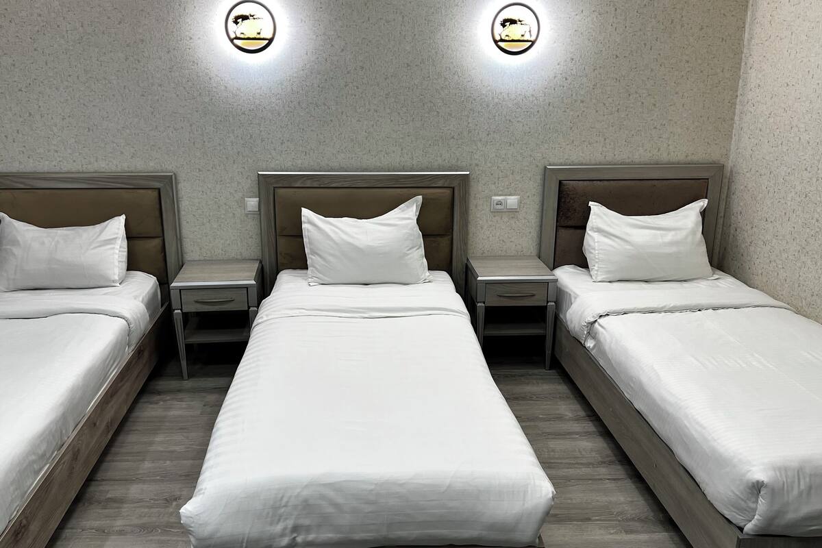 A hotel room with three single beds, each with a wooden headboard and white bedding. There are two wall-mounted light fixtures with a circular design.