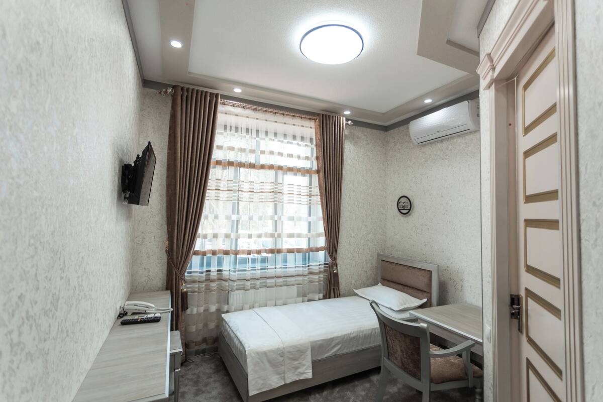 A compact hotel room with a single bed, a small desk with a chair, a flat-screen TV mounted on the wall, and a window with curtains.