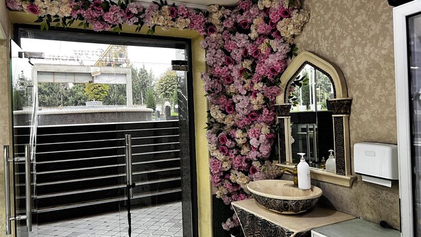 Un baño con puerta negra, un lavabo con una pileta decorativa, un espejo con un marco ornamentado y una pared adornada con flores rosadas y blancas.