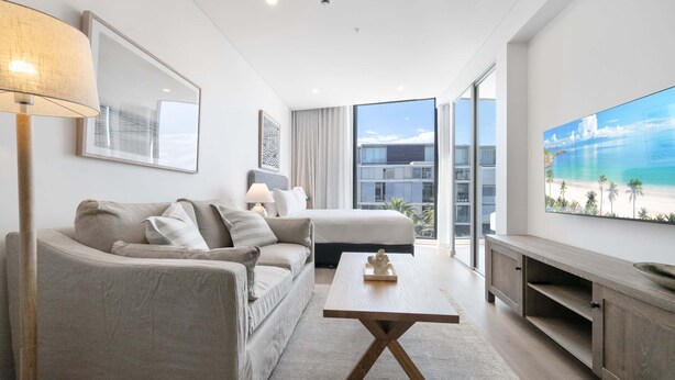 A modern living room with a sofa, a coffee table, and a TV. There is a large window with a view of buildings and a painting of a beach. - Bright Studio With Rooftop BBQ in Fremantle (Perth)