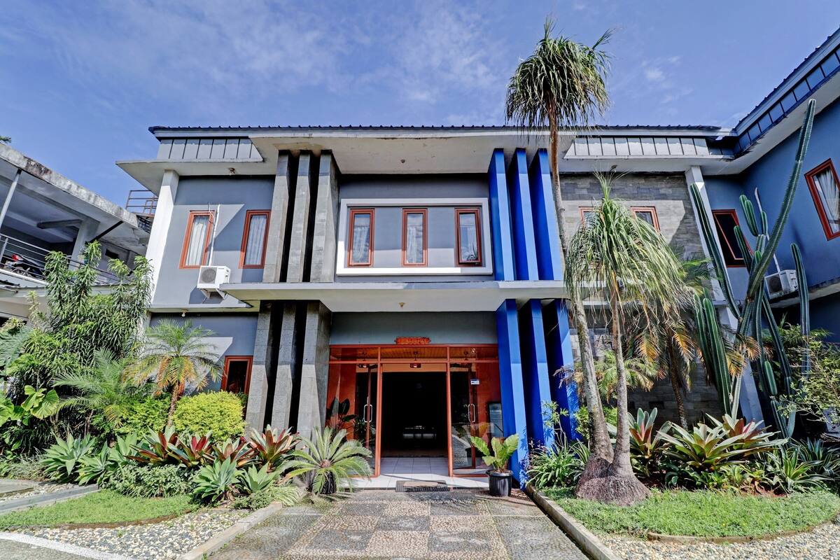 Un bâtiment moderne avec une entrée en verre, entouré de plantes tropicales et bordé d’un chemin de gravier.