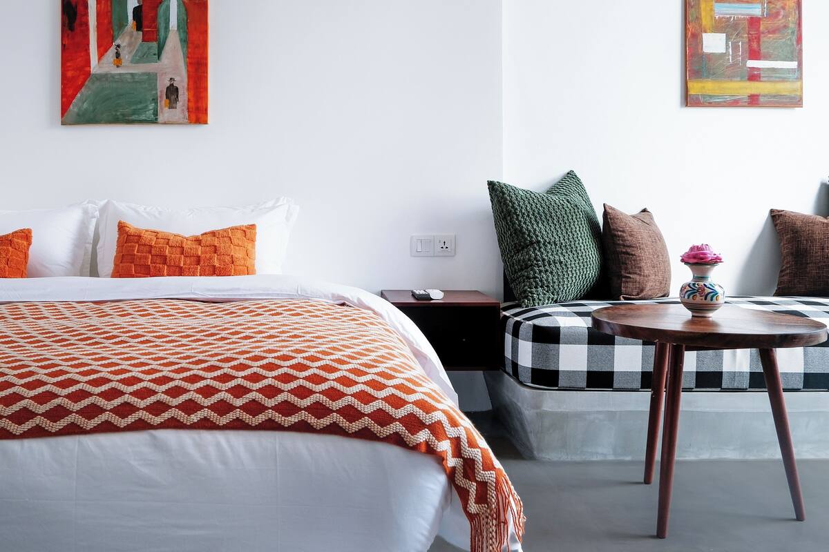 A modern hotel room with a large bed, a small sofa, a round wooden table, and two paintings on the walls.