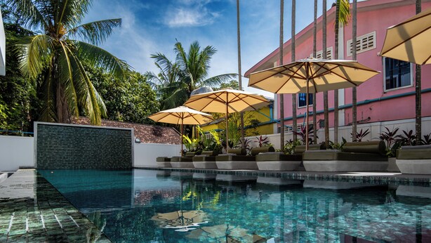 A swimming pool with clear water, surrounded by palm trees and a pink building with outdoor seating and umbrellas. - Serenity Home (Siem Reap)