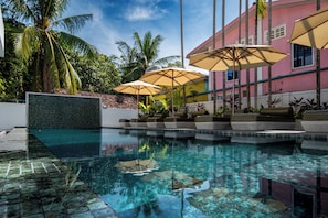 A swimming pool with clear water, surrounded by palm trees and a pink building with outdoor seating and umbrellas. - Serenity Home (Siem Reap)