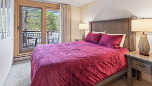 A bedroom with a large bed, a nightstand, two lamps, a window with a view of trees, and a balcony with outdoor furniture. - Best in Breck. Ski in, ski out. Pool and hot tubs. (Breckenridge)