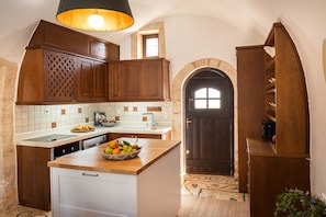 A kitchen with wooden cabinets, a central island with fruit, a built-in dishwasher, and a dark wooden door. - Tzivaeri, Lovely village house with ample outdoor area and plunge pool. (Chania)