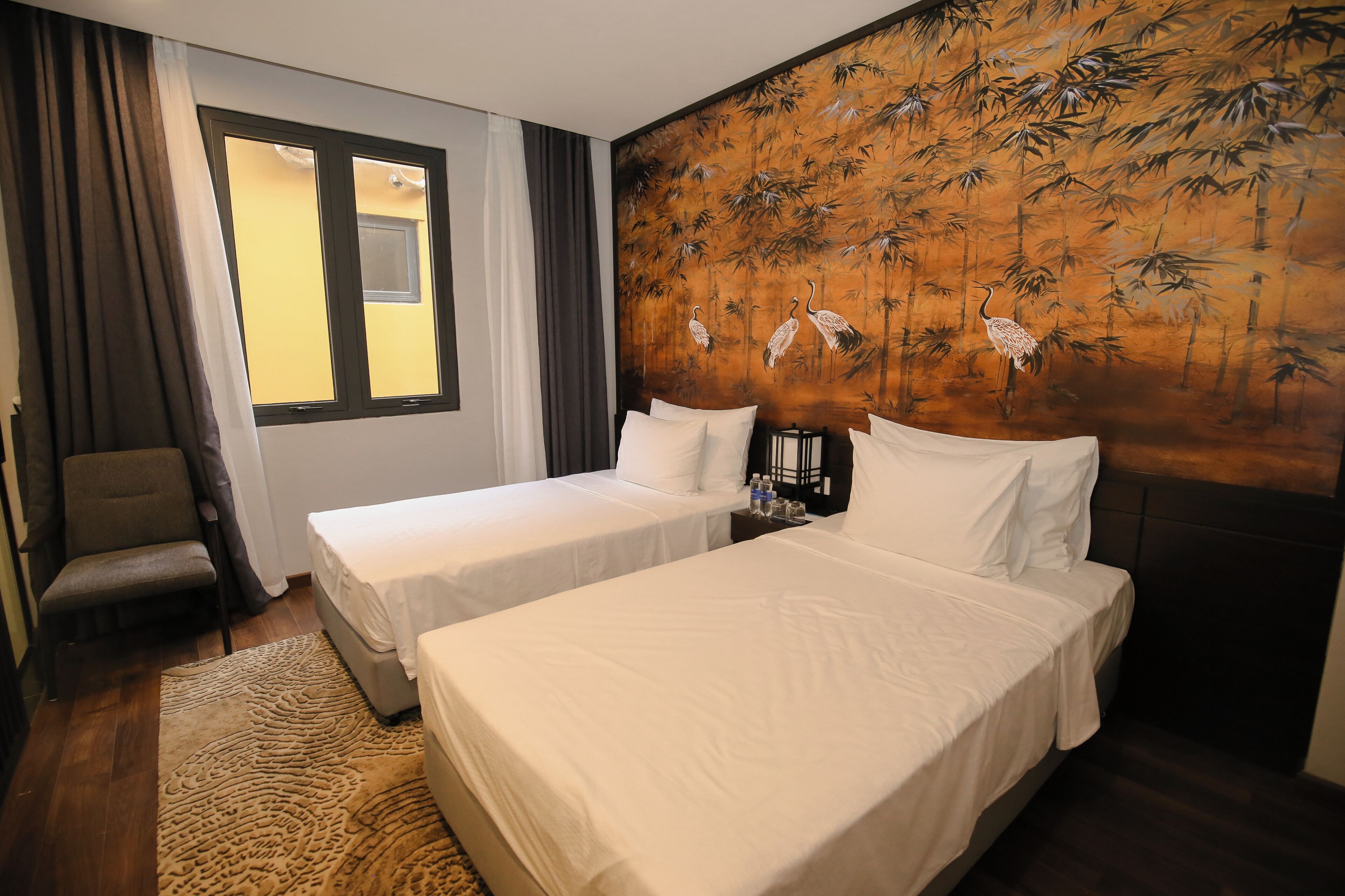 A hotel room with two single beds, a chair, a large window with curtains, and a wall mural of birds and bamboo.