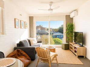 A modern living room with a sofa, a wicker chair, a television, and a large window with a view of buildings. - South Beach Sands - close to the beach (South Fremantle)