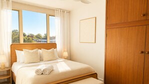 Quarto com cama de madeira, um banquinho, mesa de cabeceira e guarda-roupa.