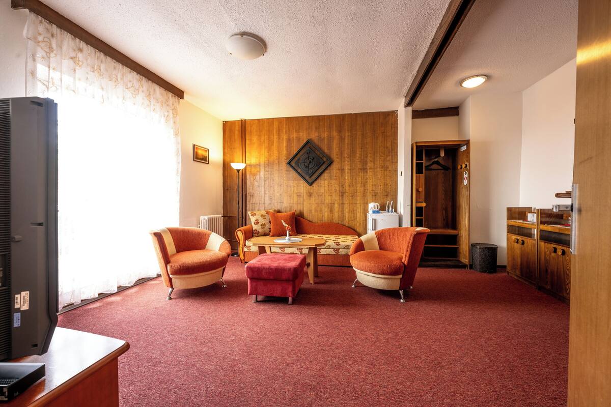A hotel room with a wooden wall, a sofa, two armchairs, a small table, a TV, and a wooden cabinet.