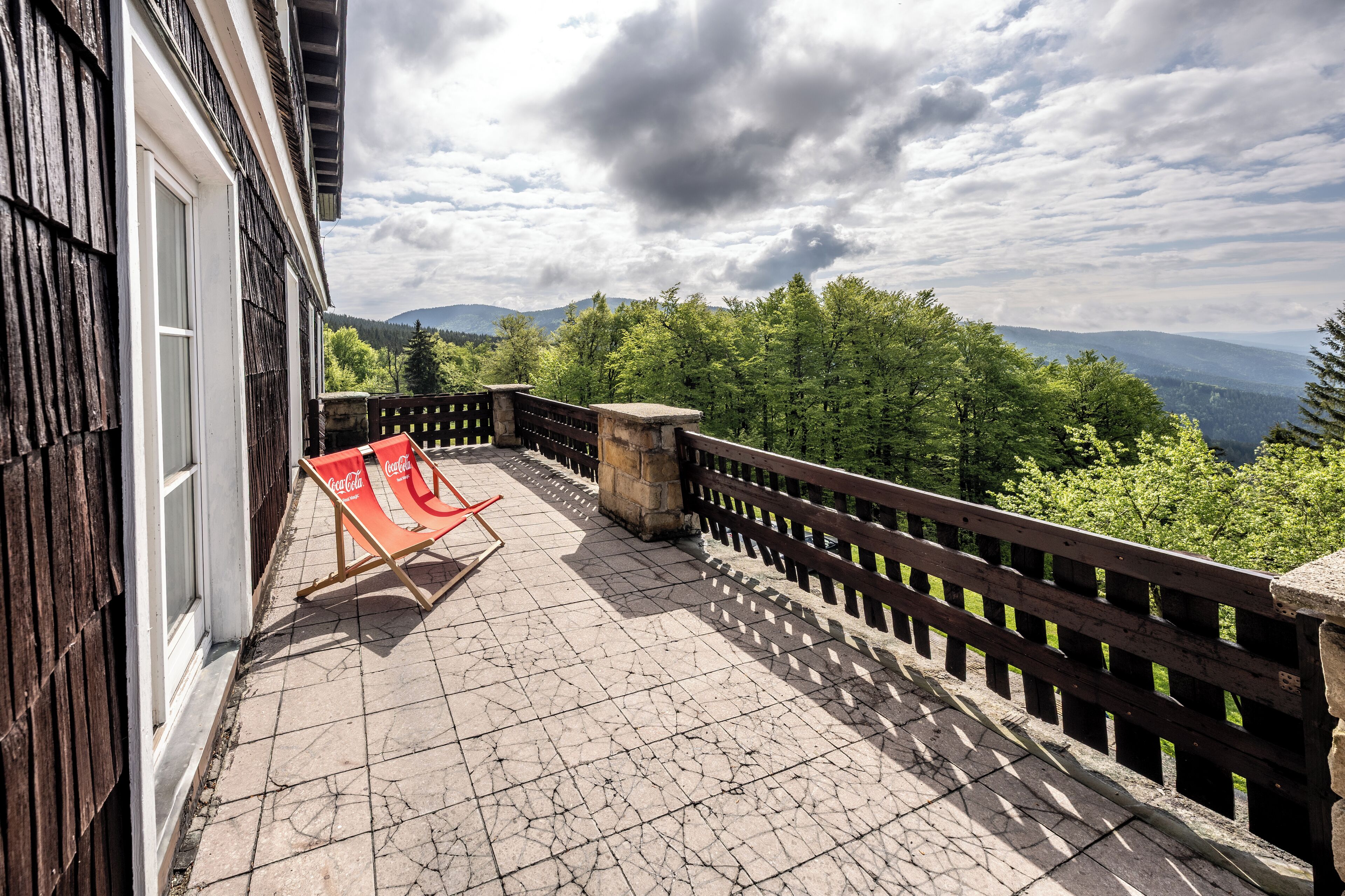 Un balcón con una silla con la marca Coca-Cola, una jardinera de piedra y barandilla de madera con vista a un paisaje boscoso.