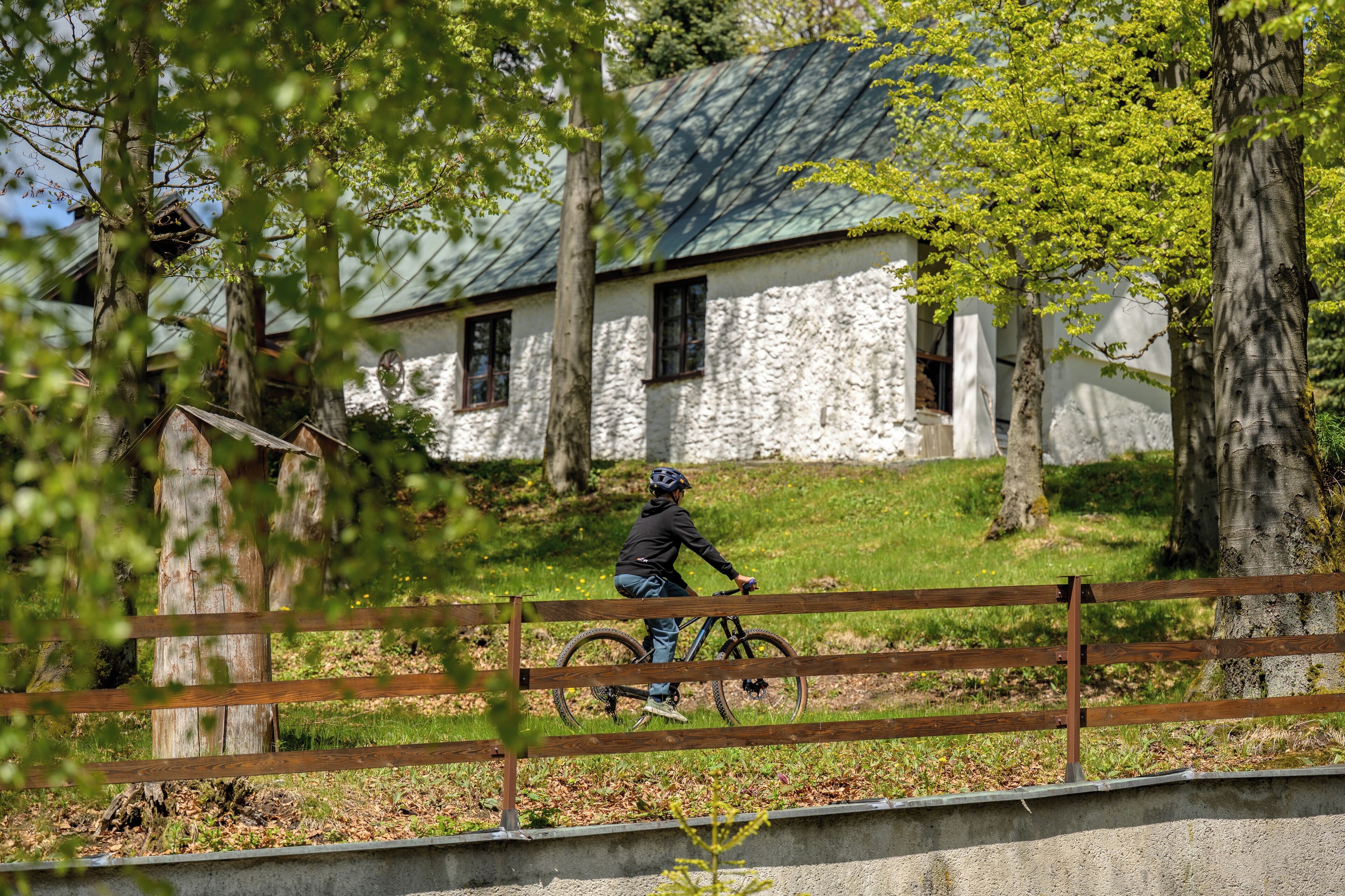 A person riding a bicycle near a stone building with a green roof, surrounded by trees.