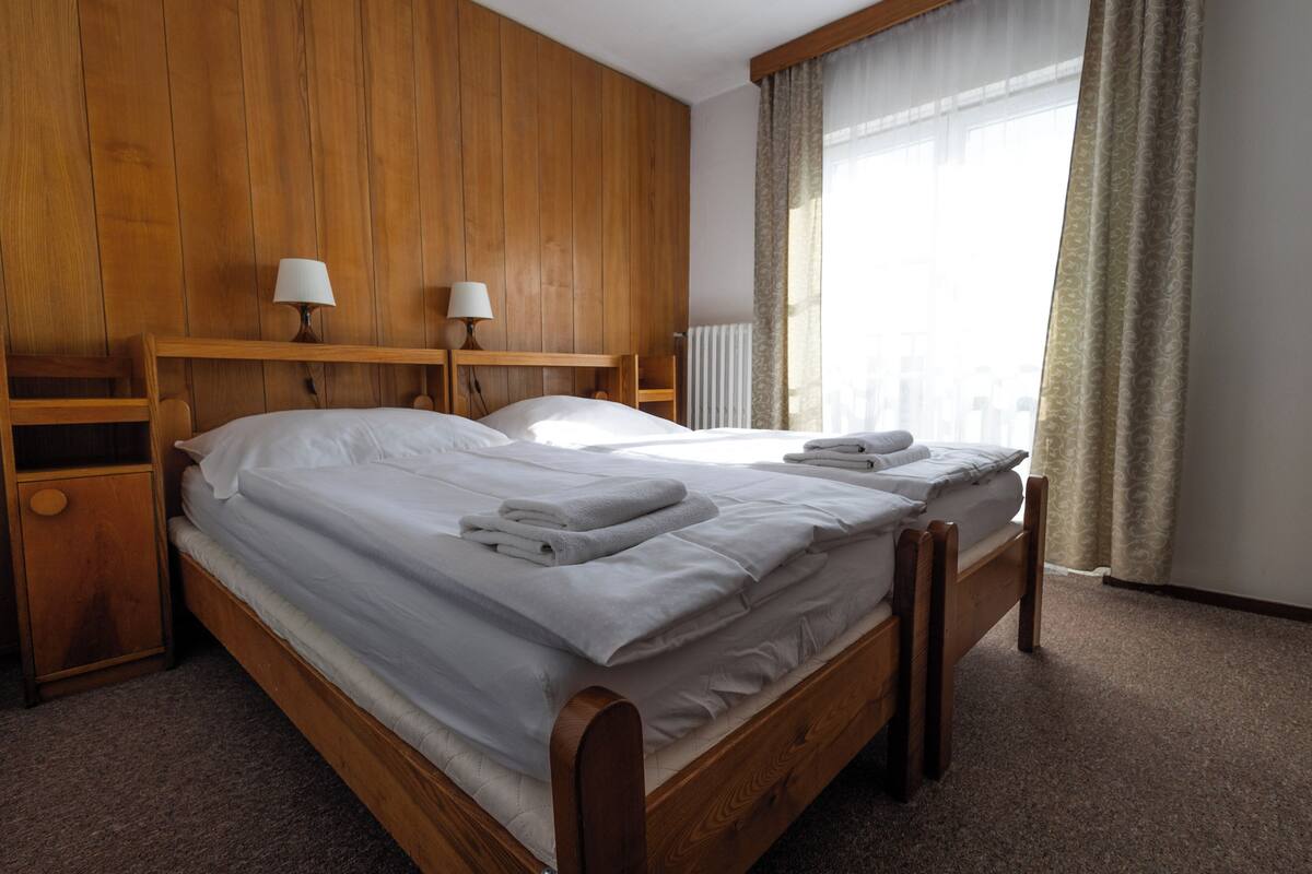 A wooden bed with white linens and towels, a nightstand with a lamp, and a window with curtains.