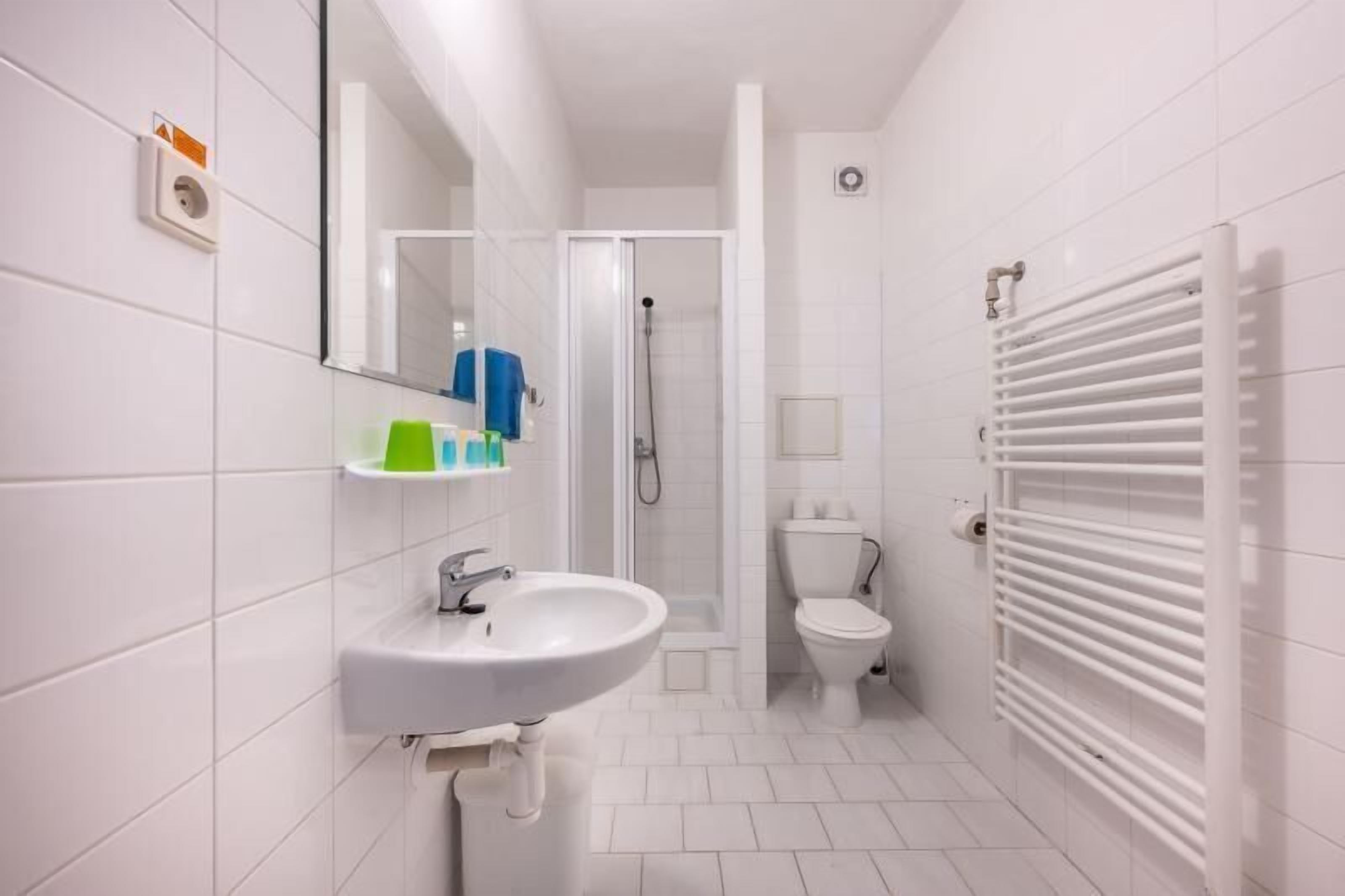 Casa de banho compacta com lavatório branco, sanita, chuveiro e porta-toalhas.