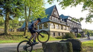 A historic building with a steeply pitched roof and large windows, situated next to a paved path where cyclists are riding. - Hotel Tanečnica (Prostredni Becva)