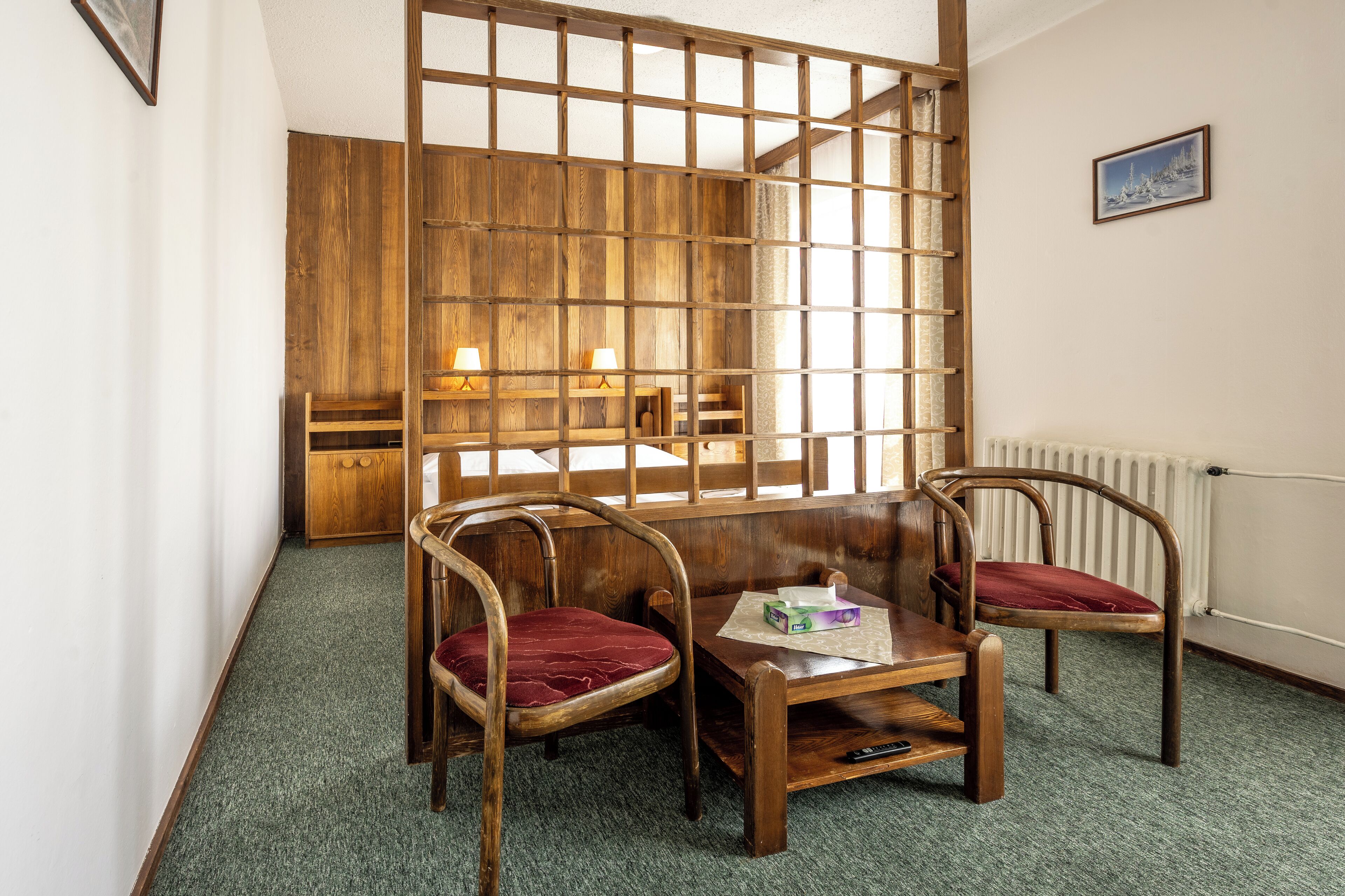 Ein Zimmer mit einer Holzwand, zwei Holzstühle mit roten Kissen, ein kleiner Holztisch mit einer Schachtel und Fernbedienung sowie ein Heizkörper an der Wand.