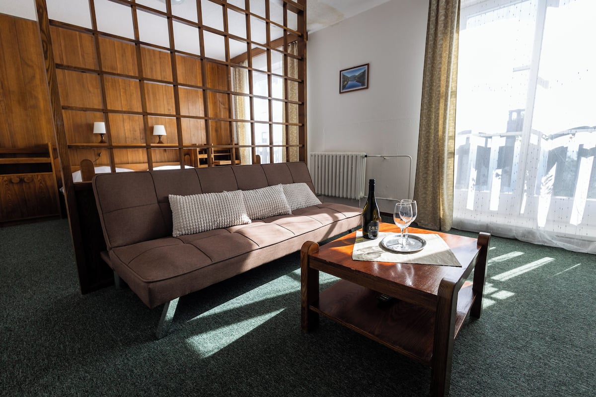 A room with a brown sofa, a wooden coffee table with a bottle and glass, and a view of the outside through a window.