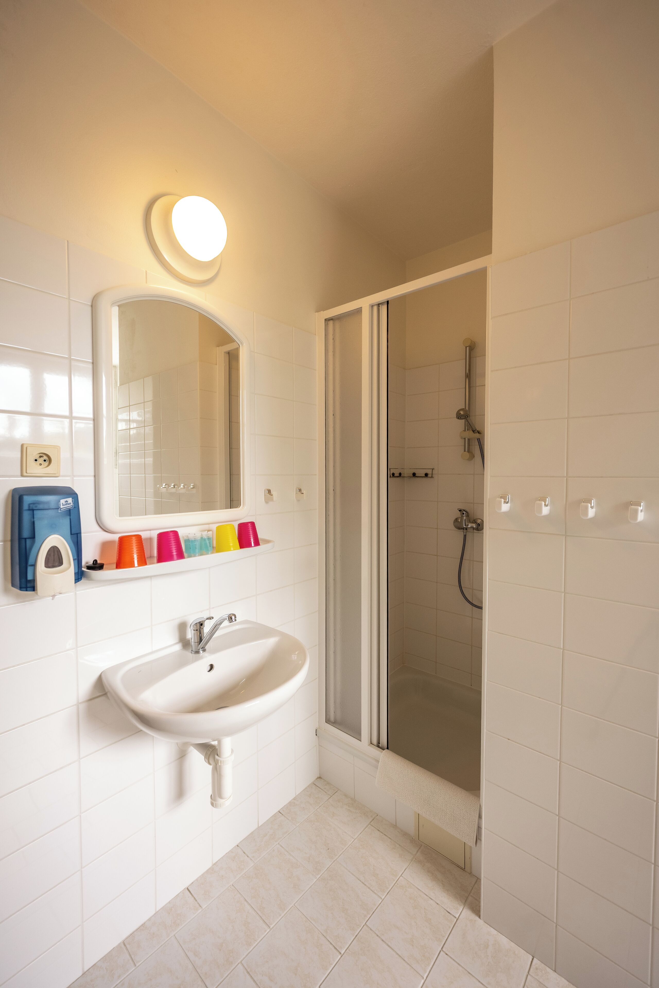 Ein Badezimmer mit einem weißen Waschbecken, einem Spiegel, einer Dusche und einem Regal mit bunten Tassen.