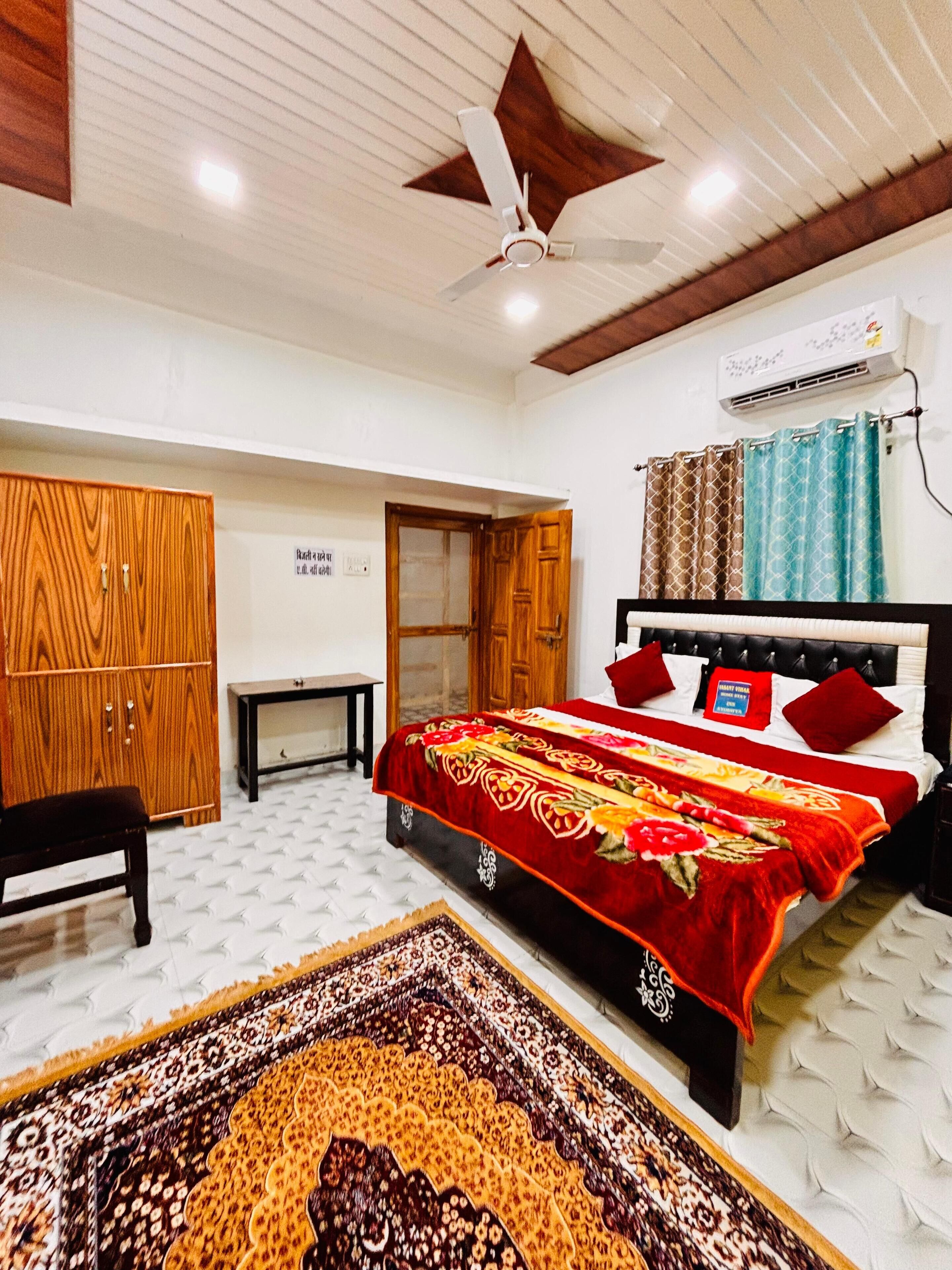 A bedroom with a bed, a wardrobe, a small table, a ceiling fan, and an air conditioning unit.