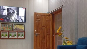 A living room with a blue sofa, a glass coffee table, a wooden door, and a TV mounted on the wall. - Vasant Vihar Inn (Ayodhya)