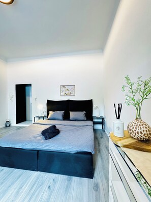 A modern bedroom with a large bed, a nightstand, a vase with flowers, and a wall-mounted artwork.