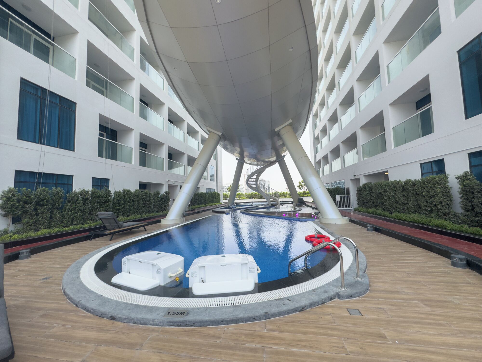 A modern indoor pool with a circular design, surrounded by a wooden deck and a white building with balconies.