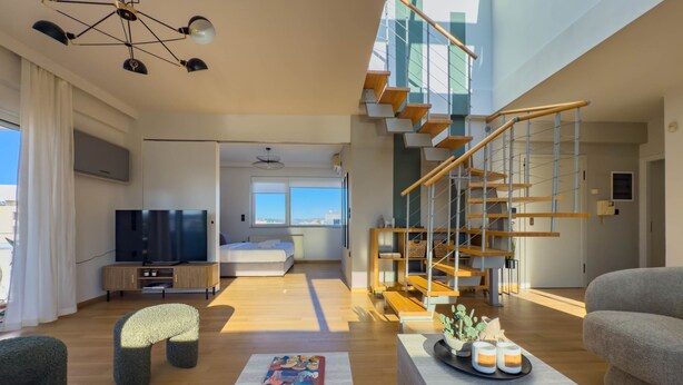 A modern living room with a large TV, comfortable seating, a staircase, and a view to the outside. - Acropolis Sky Retreat Jacuzzi 3BR Maisonette (Athens)