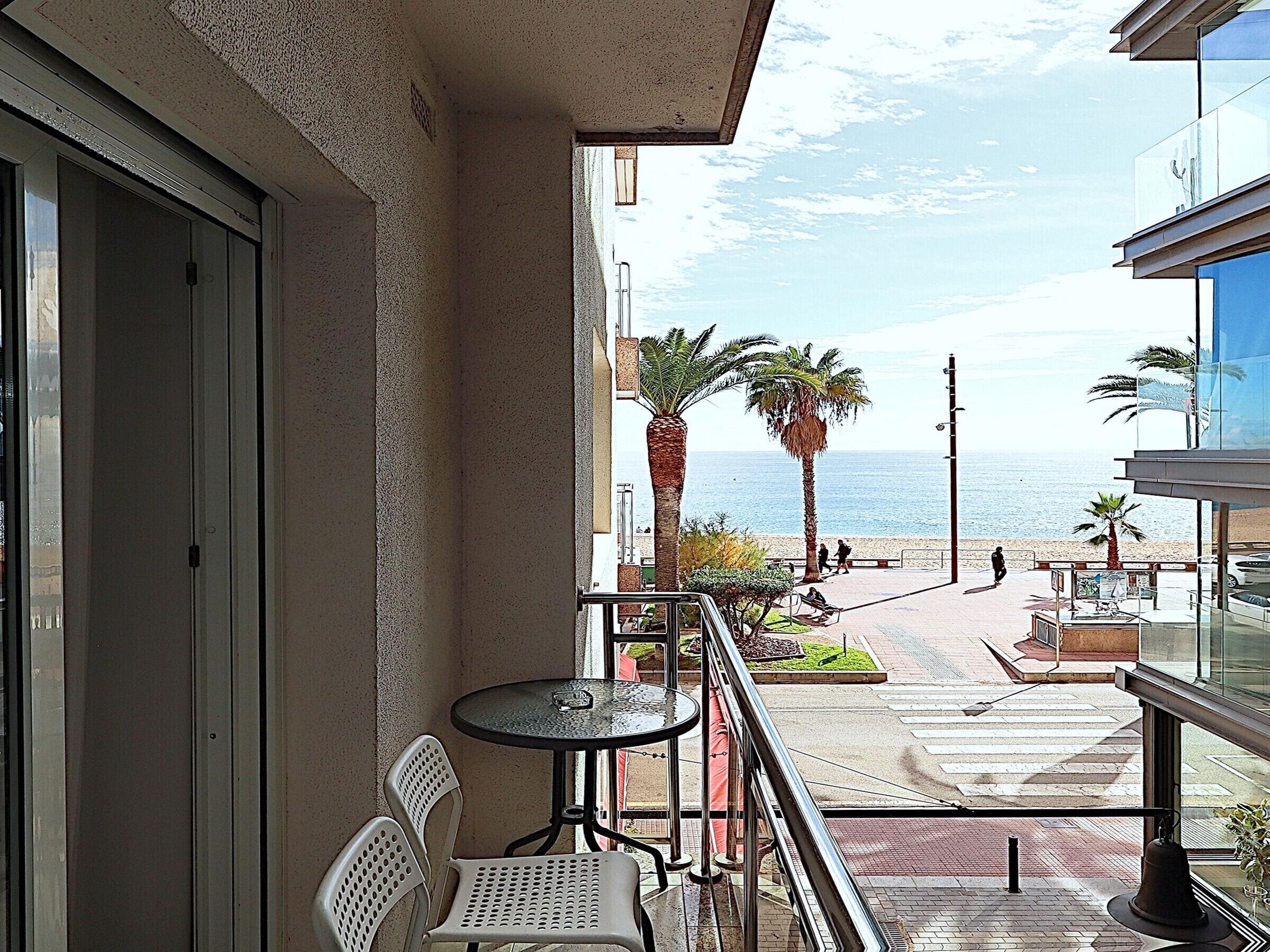 En balkong med ett bord och stolar, med utsikt över en strand med palmer och en klar himmel.