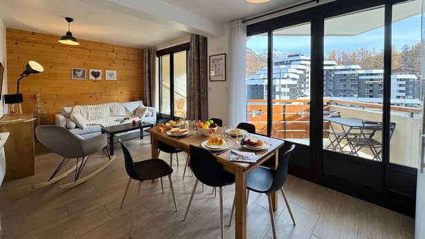 Una sala de estar moderna con una pared de madera, un comedor con una mesa dispuesta para dos personas, y amplias ventanas que ofrecen vistas a edificios y árboles.