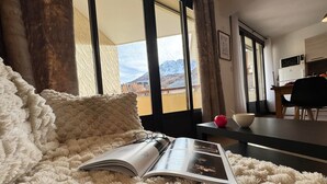 Una habitación acogedora con un ventanal grande, una alfombra blanca y mullida, una mesa con un bol y una manzana, y una revista sobre la alfombra.