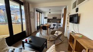 Una sala de estar moderna con un sofá, una mesa de centro y un comedor con mesa y sillas. Se ve una cocina al fondo con armarios y un televisor montado en la pared.