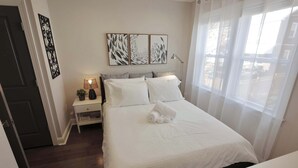 A neatly made bed with white linens and rolled towels, a bedside table with a lamp, and a window with sheer curtains. - The Brightwood House - Family Living in the Heart of DC! (Washington)