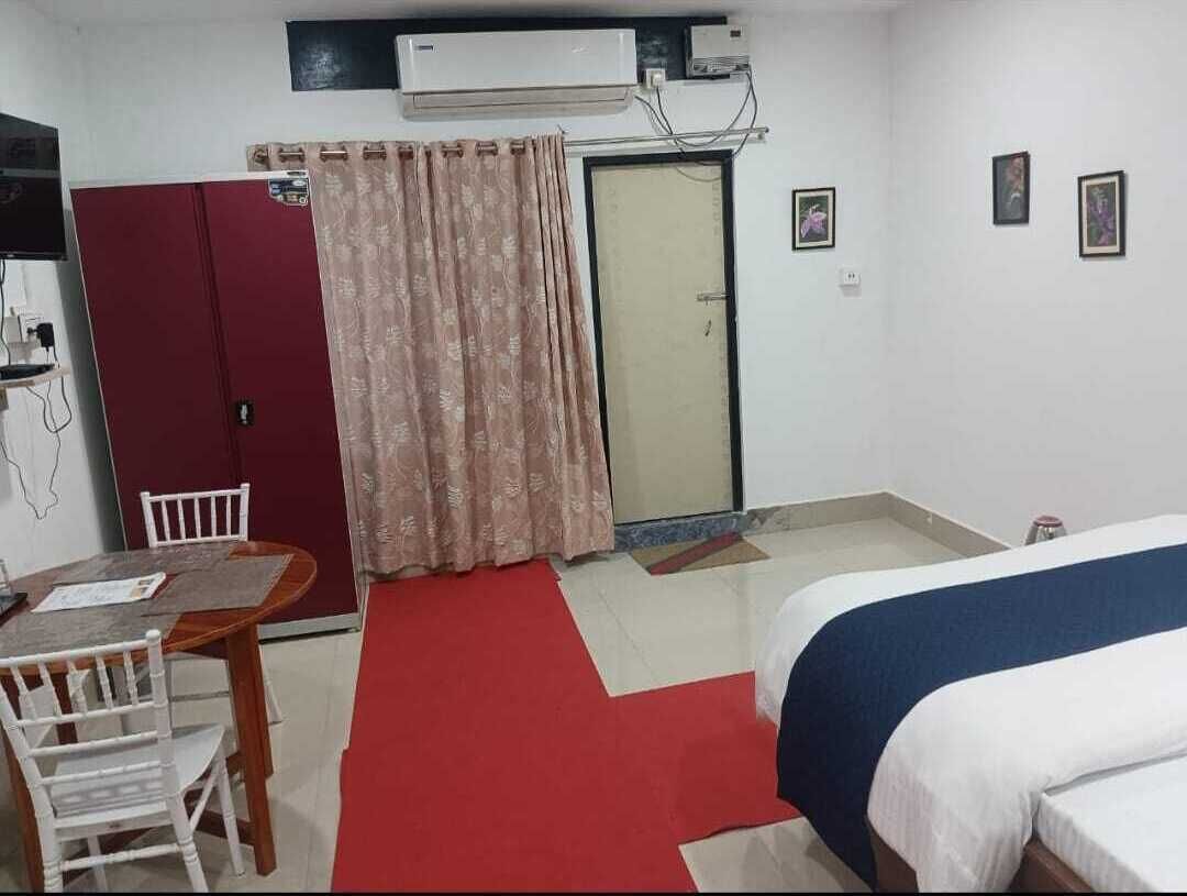 Una habitación de hotel con cama, un escritorio con sillas, una alfombra roja, un armario, una puerta y un equipo de aire acondicionado.