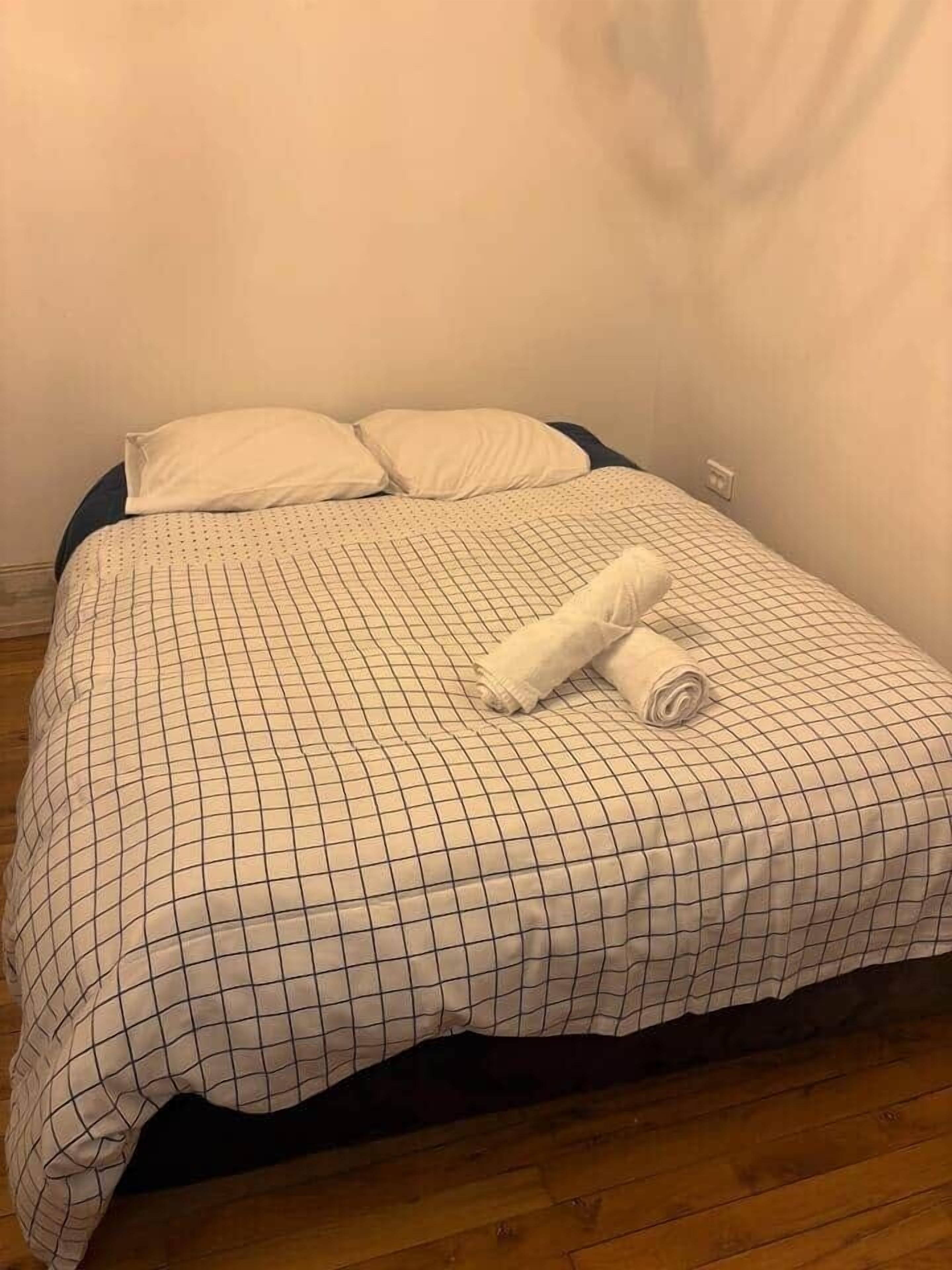 Un letto rifatto con una coperta a quadri e due cuscini, un asciugamano arrotolato sul letto, e un pavimento in legno.