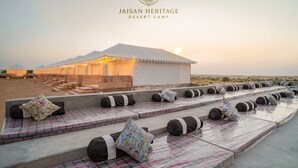 Un campamento en el desierto con una estructura tipo tienda, asientos al aire libre con cojines y un paisaje desértico al fondo.