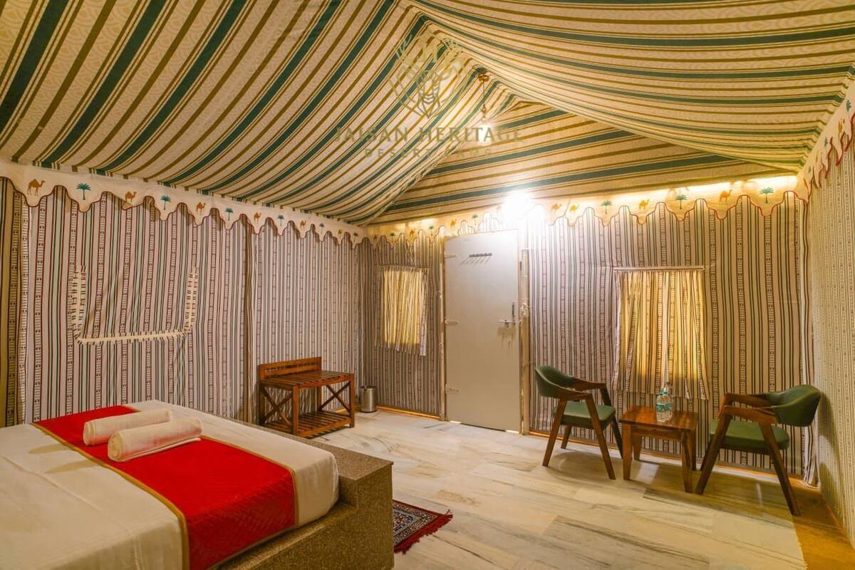 Una habitación con cortinas a rayas, una cama con colcha roja, una silla de madera, una mesita y una puerta.