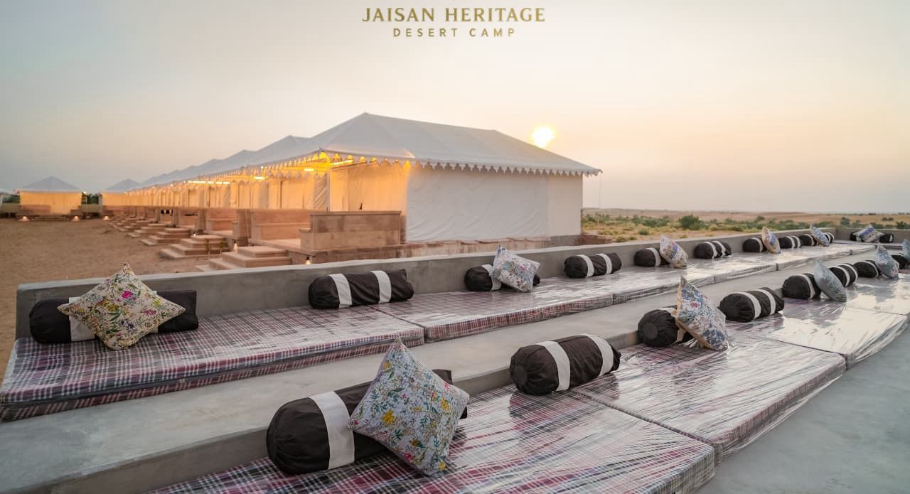 Un campamento en el desierto con una estructura tipo tienda, asientos al aire libre con cojines y un paisaje desértico al fondo.