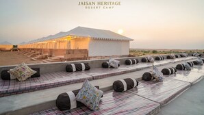 Un campamento en el desierto con una estructura tipo tienda, asientos al aire libre con cojines y un paisaje desértico al fondo.