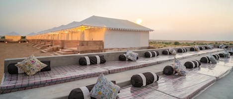 A desert camp with a tented structure, outdoor seating with cushions, and a desert landscape in the background.