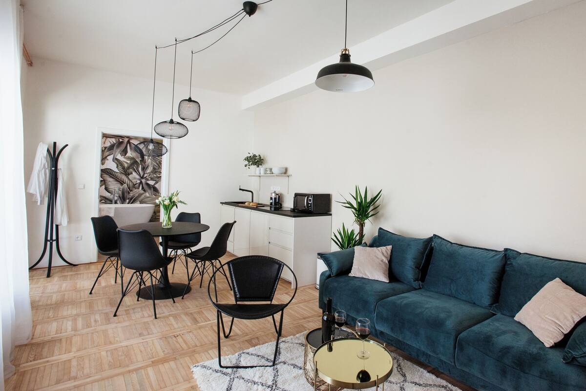 A modern living room with a dark green sofa, black chairs, a dining table, and a kitchen area in the background.