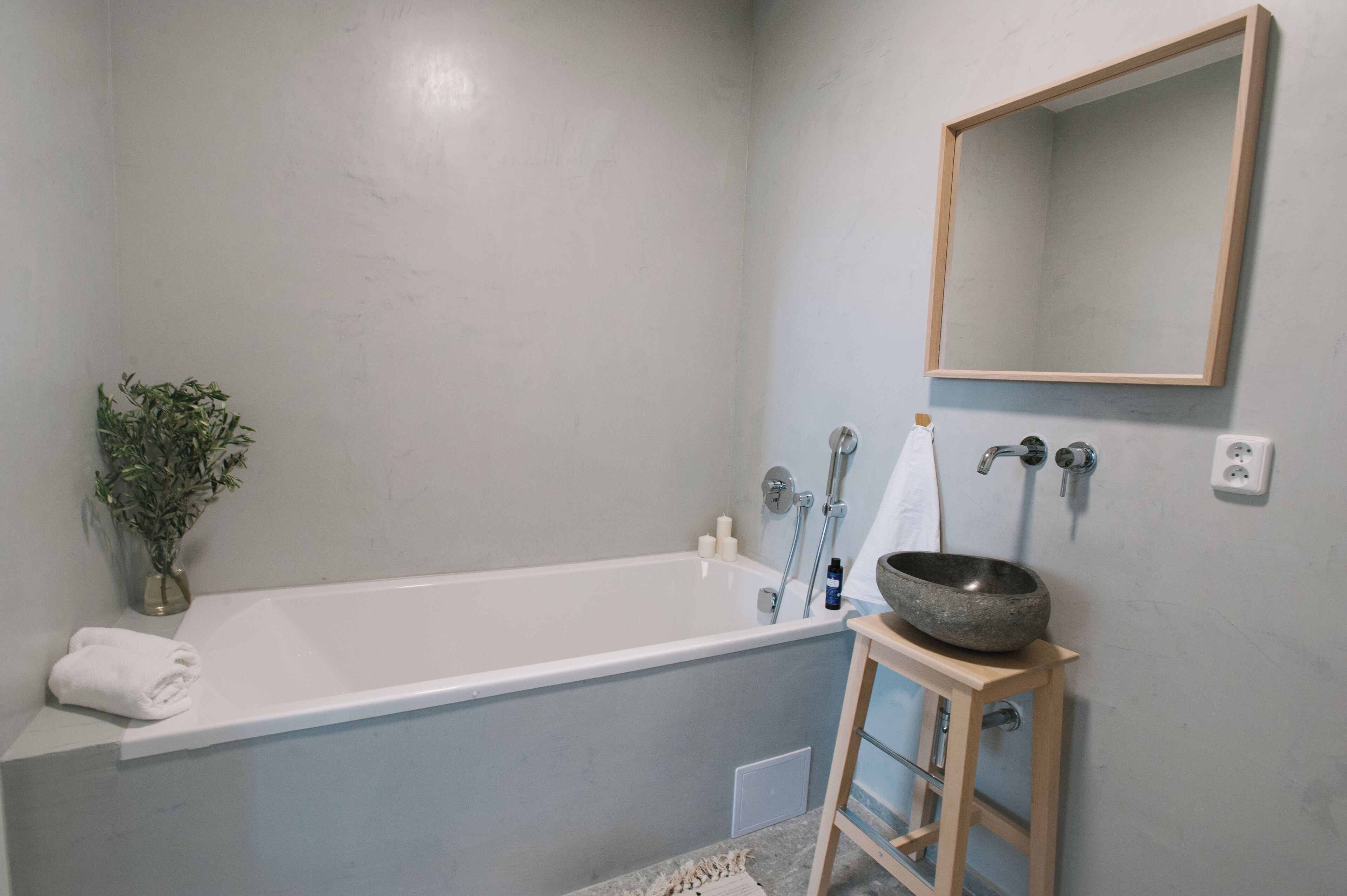 A bathroom with a white bathtub, a wooden stool with a stone basin, a potted plant, and a wall-mounted mirror.