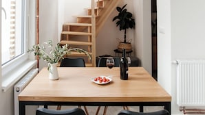 Een eethoek met een houten tafel, zwarte stoelen, een potplant, een fles wijn en een bord eten.