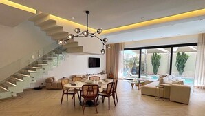 A spacious living area with a modern staircase, a dining table with chairs, a sofa, and a large window with a view of a pool and greenery. - Palais Oyena (Marrakech)